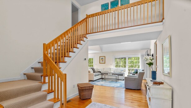 Un escalier avec des rampes en bois menant à un niveau supérieur, un salon avec un canapé et une table basse, et une vue sur l’extérieur grâce à de grandes fenêtres.