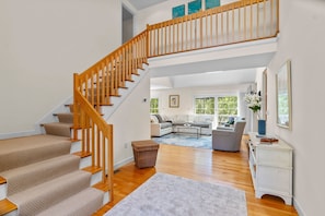 Un escalier avec des rampes en bois menant à un niveau supérieur, un salon avec un canapé et une table basse, et une vue sur l’extérieur grâce à de grandes fenêtres.