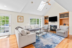 A spacious living room with a large sofa, a coffee table, and a fireplace. There are large windows, a ceiling fan, and a flat-screen TV mounted on the wall. - New Rental-Bright Coves, Walk to Marina and Popponesset Spit and clean! (Mashpee)