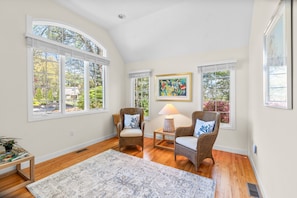 A room with a high ceiling, large windows, wicker chairs, a rug, and a painting on the wall.
