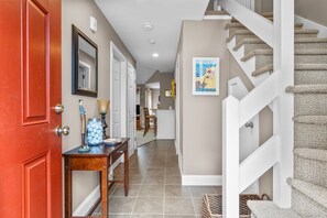 A hallway with a red door, a wooden table with a jar of balls, a framed mirror, and a staircase with a textured carpet.