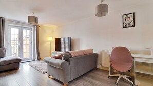 Ein modernes Wohnzimmer mit einer Couch, einem Schreibtisch, einem Sessel, einem Fernseher und einem Fenster mit Vorhängen.