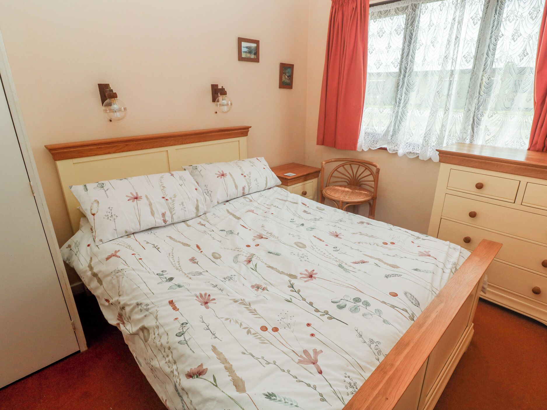 Un dormitorio con colcha de estampado floral, cabecero de madera, silla de mimbre y ventana con cortinas de encaje.