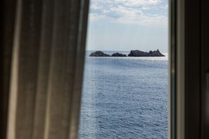 Blick durch ein Fenster mit Vorhang auf ein ruhiges Meer und entfernte Felsen.