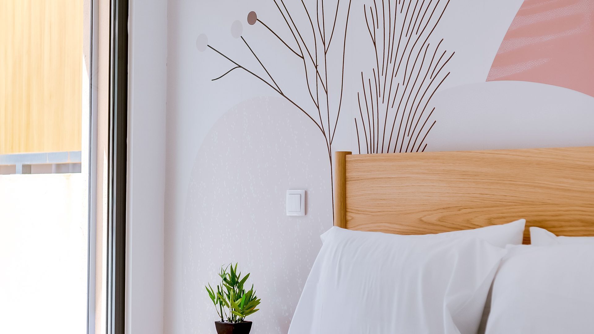 A bedroom with a wooden headboard, white bedding, a small potted plant, and a wall-mounted light switch.