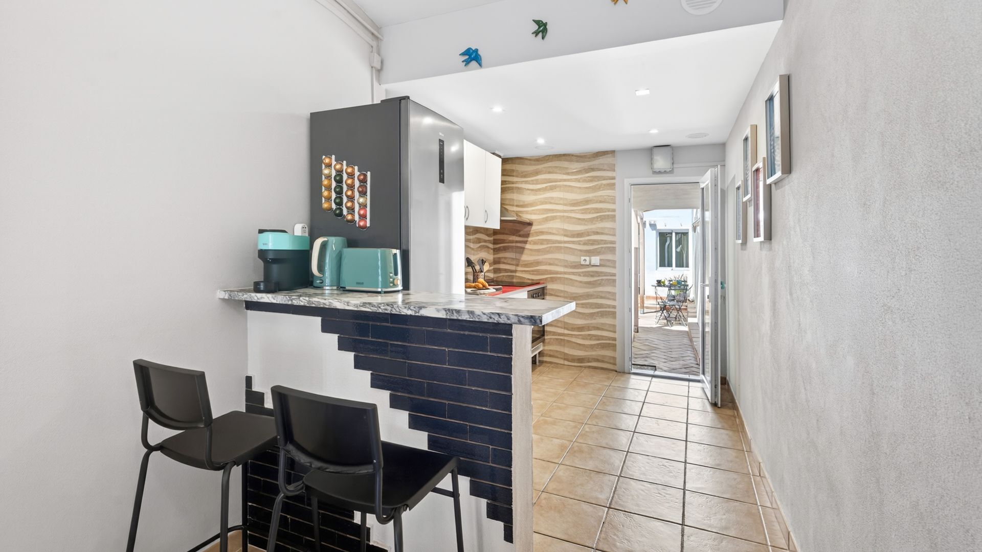 Une cuisine moderne avec un plan de travail en marbre, deux chaises noires, un réfrigérateur et un élément décoratif mural.