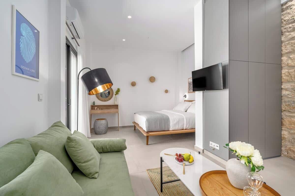 Une chambre d’hôtel moderne avec un canapé vert, un lit avec une literie blanche, un téléviseur fixé au mur et une petite table sur laquelle est posé un vase de fleurs.