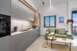 Eine moderne Küche mit grauen Schränken, einem schwarzen Backofen und einer Kaffeemaschine. Ein grünes Sofa mit einer Blumenvase und ein Tisch mit einer Obstschale. Ein großes Fenster bietet Blick auf die Stadt.