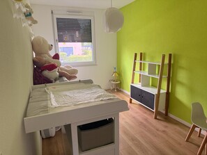 Ein Kinderzimmer mit einem Kinderbett, einem großen Teddybären, einem Regal und einem Fenster mit Jalousien.
