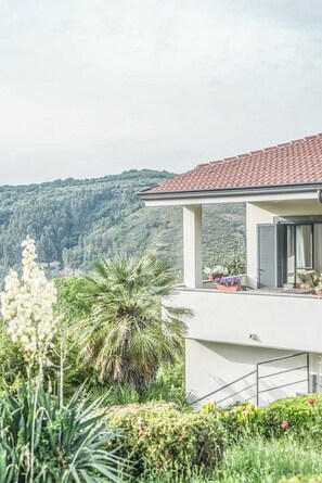 Ein modernes Haus mit rotem Ziegeldach, einem Balkon mit Topfpflanzen und Blick auf einen bewaldeten Hang.