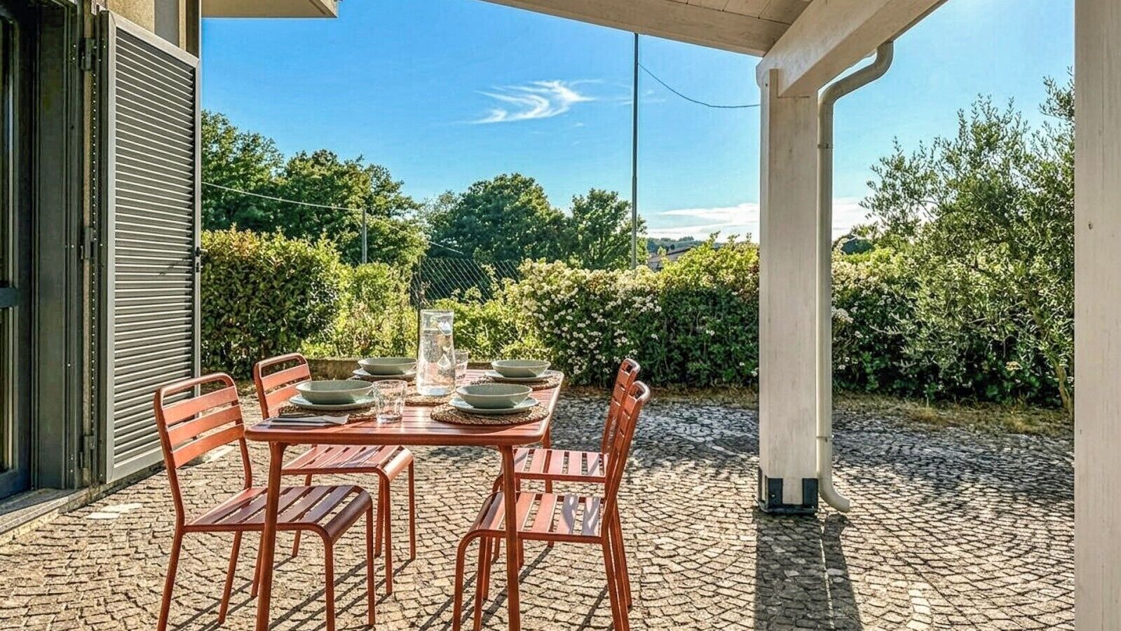 An outdoor patio with a table set for four, surrounded by greenery and a clear sky.