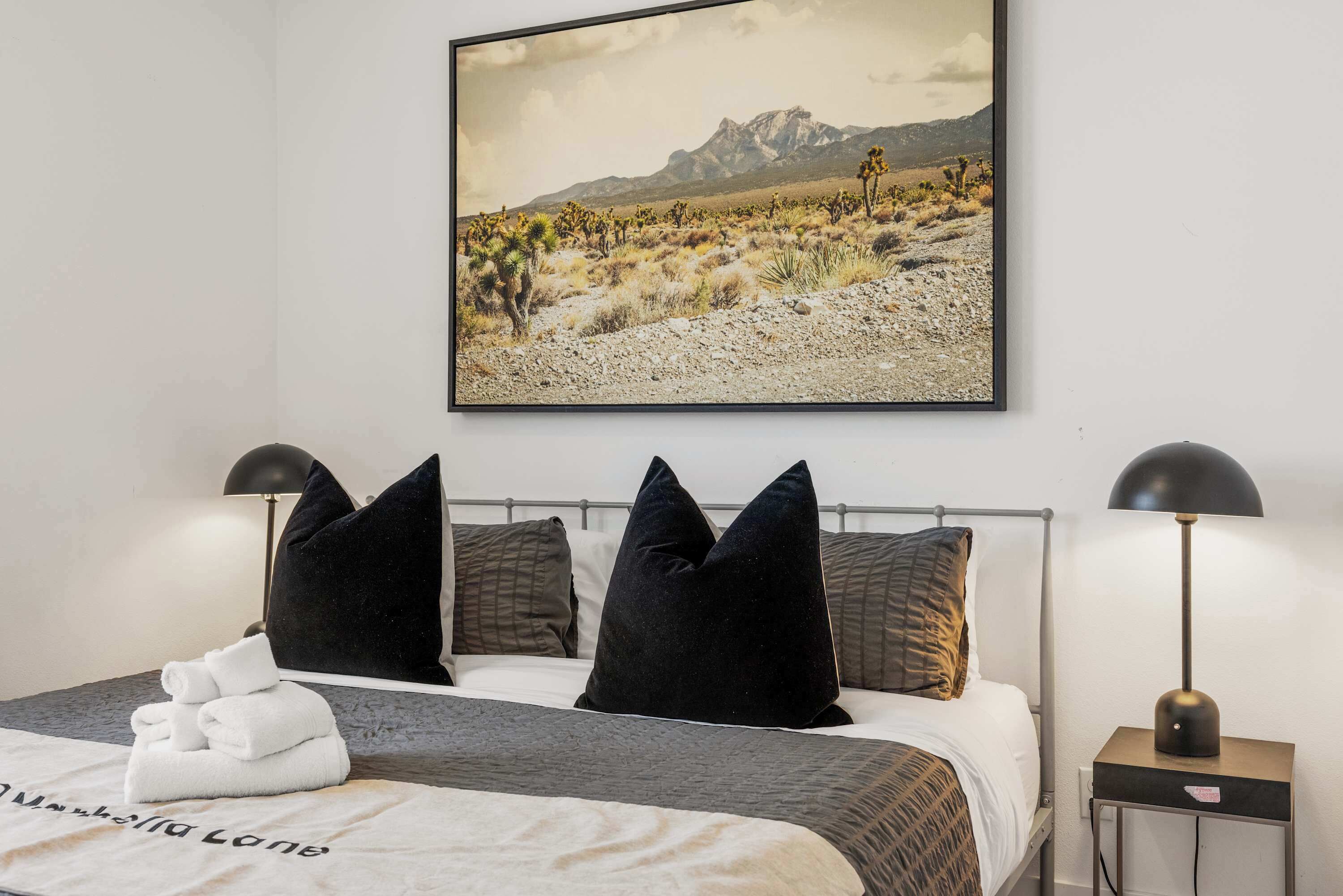 Een netjes opgemaakt bed met een witte handdoek, zwarte en bruine kussens, en een schilderij van een woestijnlandschap erboven.