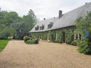 A building with a gravel driveway leading to a covered entrance, surrounded by greenery. - A country cottage located in a lush, green setting near the sea and the Seine. (Foucart)