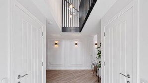 Un couloir avec des portes blanches, un plancher en bois, un escalier avec des rampes noires et des luminaires fixés au mur.