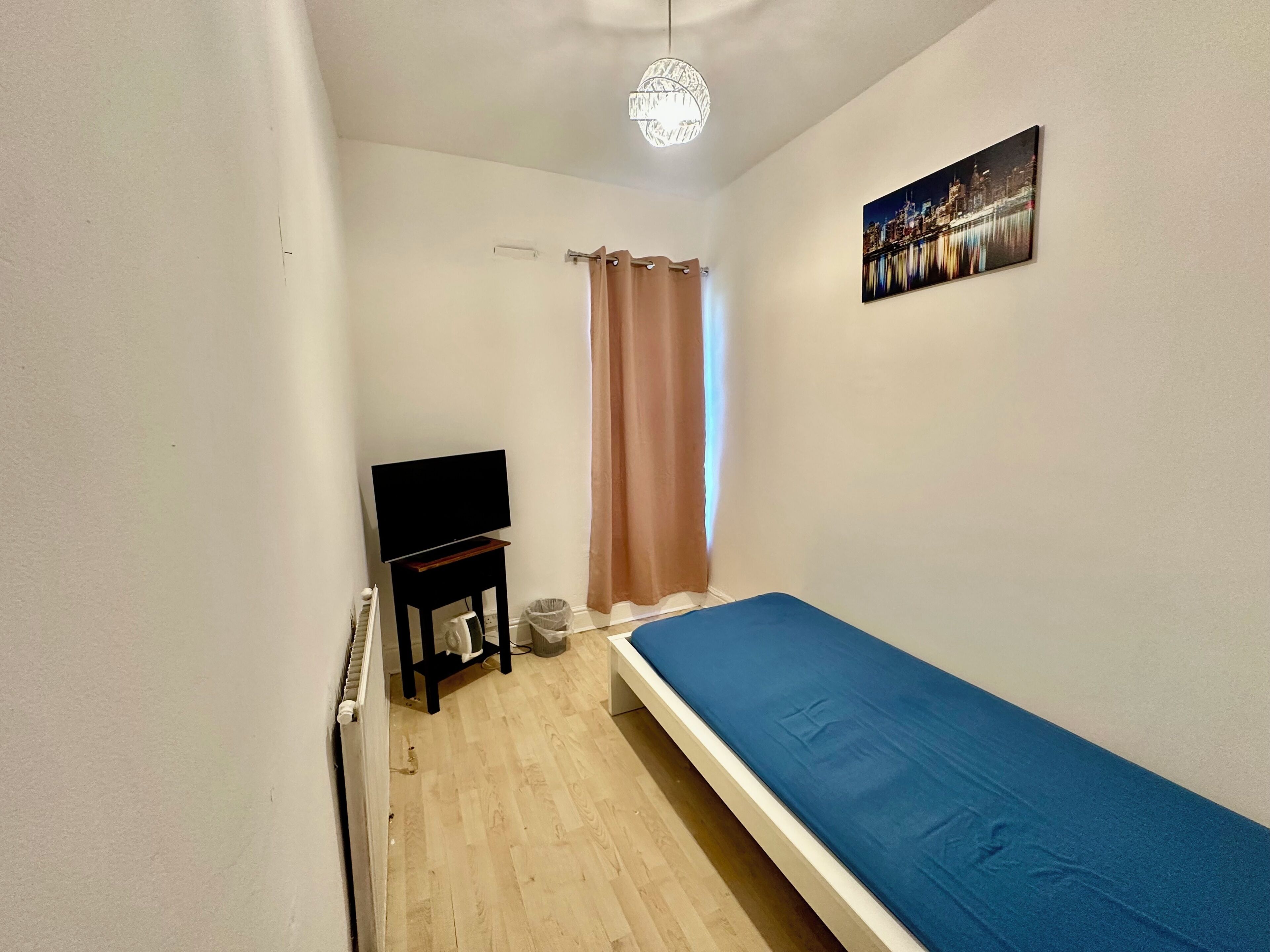 Een kleine, smalle kamer met een enkel bed, een televisie op een standaard, een radiator en een schilderij aan de muur.