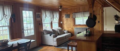 A cozy cabin interior with a wooden ceiling, a sofa, a dining table with chairs, and a kitchen area with a stove and a hanging pot.