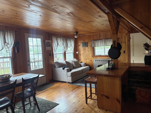 A cozy cabin interior with a wooden ceiling, a sofa, a dining table with chairs, and a kitchen area with a stove and a hanging pot.