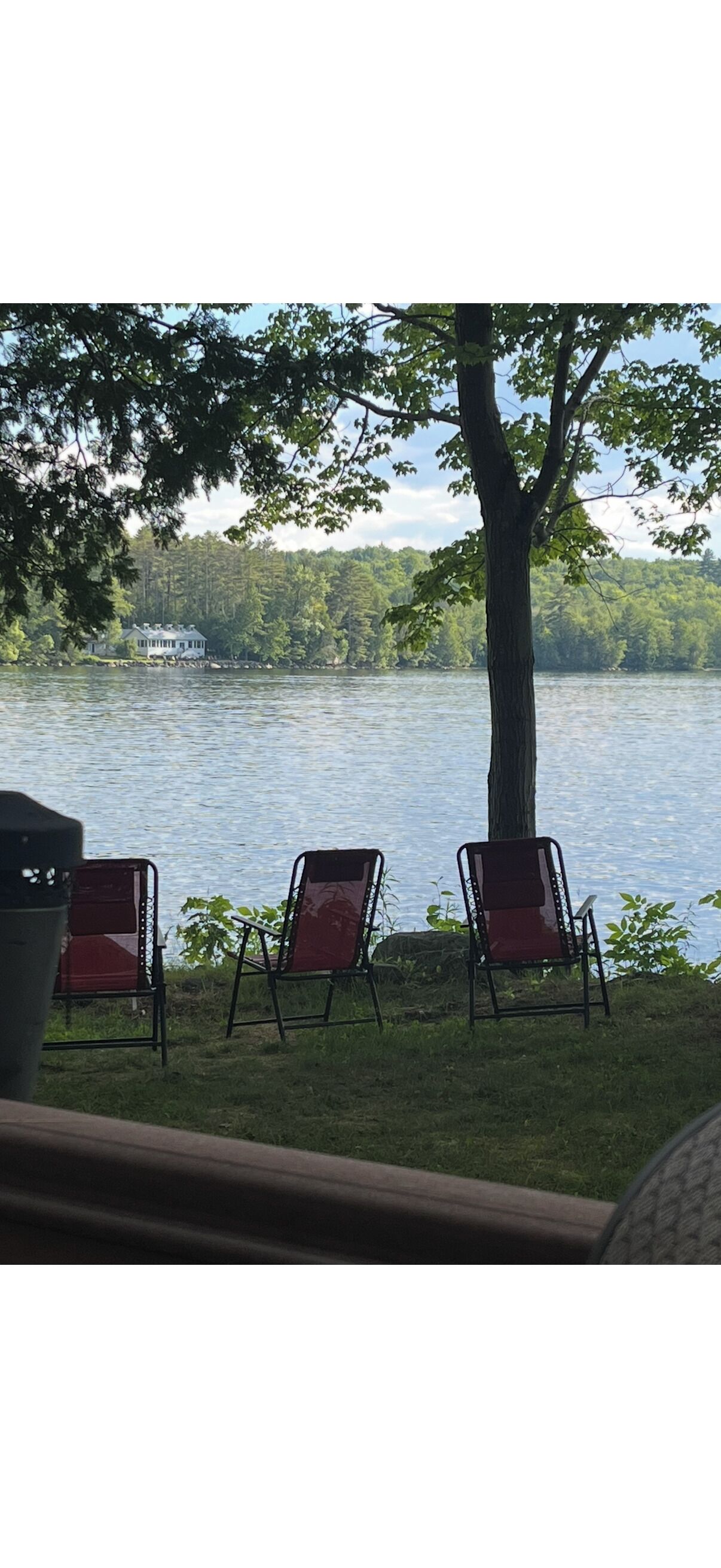 Vista sul lago con tre sedie e un albero, che si affaccia su una casa dall'altra parte dell'acqua.