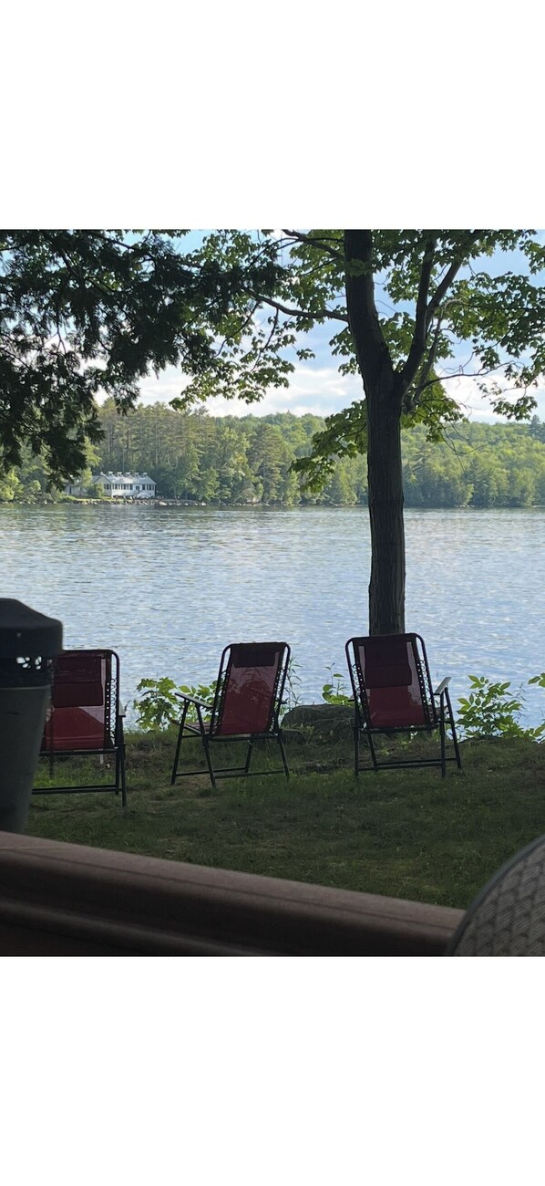 A lakeside view with three chairs and a tree, overlooking a house across the water.