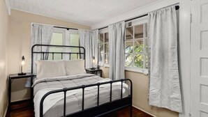 Ein Schlafzimmer mit schwarzem Metallbettgestell, zwei Nachttischen inklusive Lampen und weißen Vorhängen an den Fenstern.