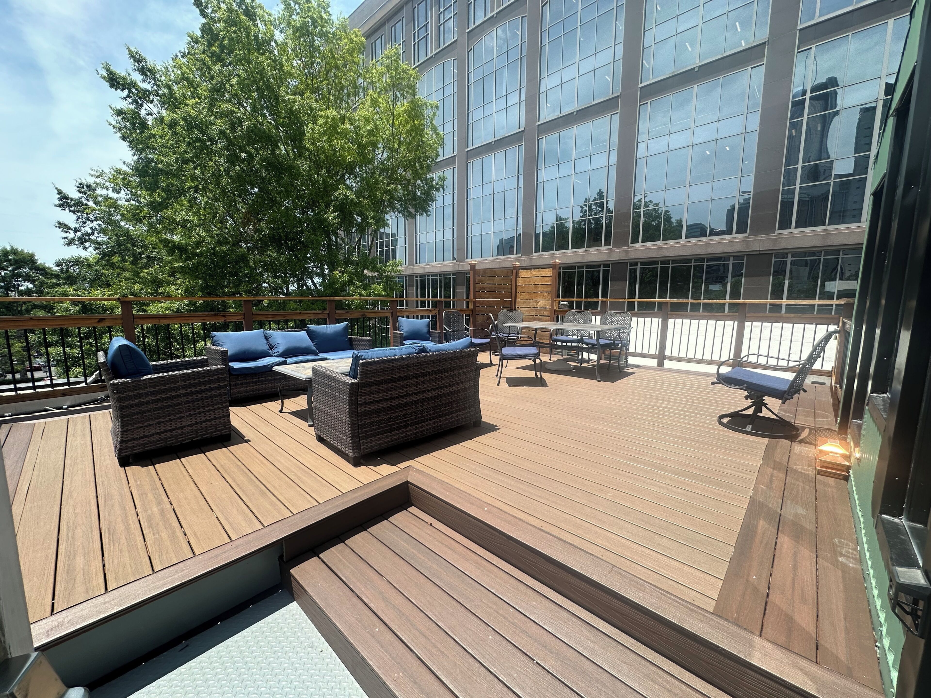 A wooden deck with outdoor furniture, a large windowed building in the background, and a tree.