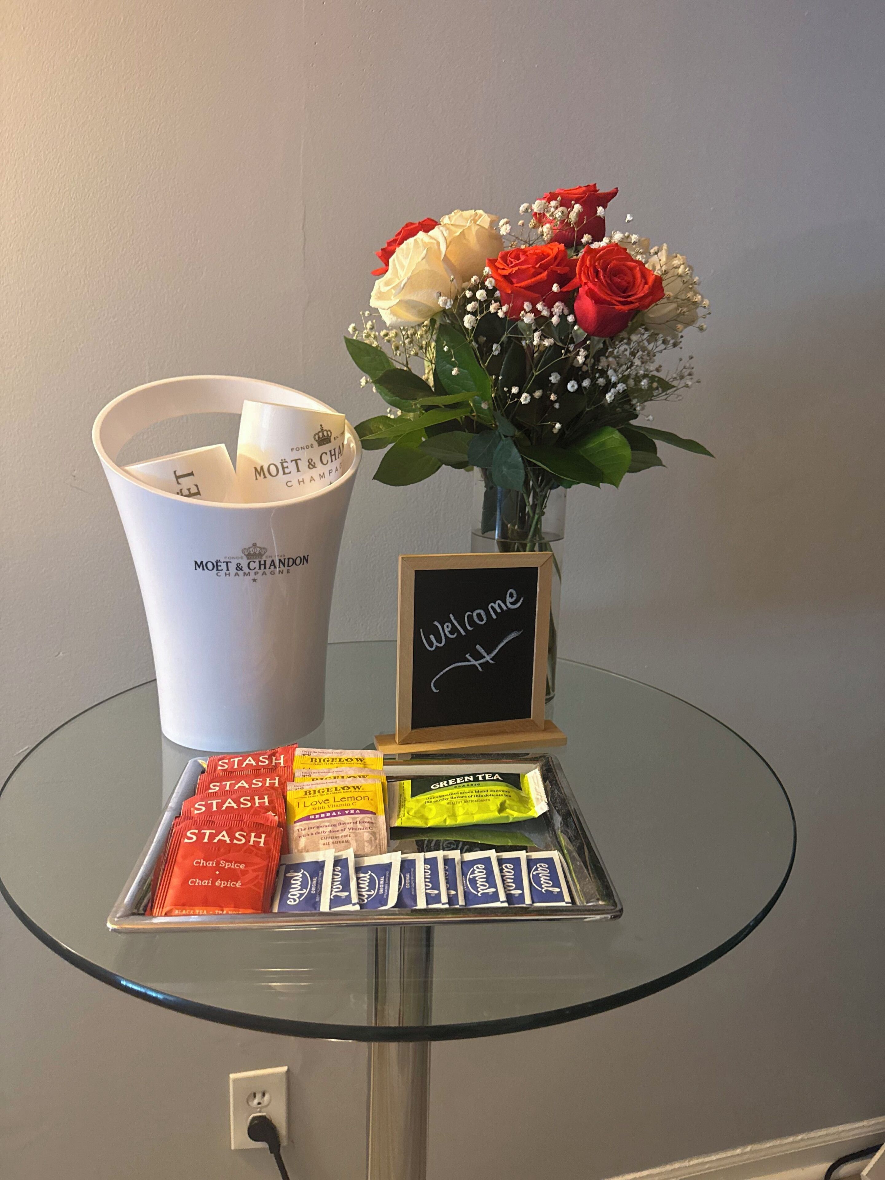 Una mesa de vidrio con un letrero de bienvenida, un ramo de flores, un balde con la etiqueta 'MOËT & CHANDON CHAMPAGNE' y varios paquetes de té y especias.