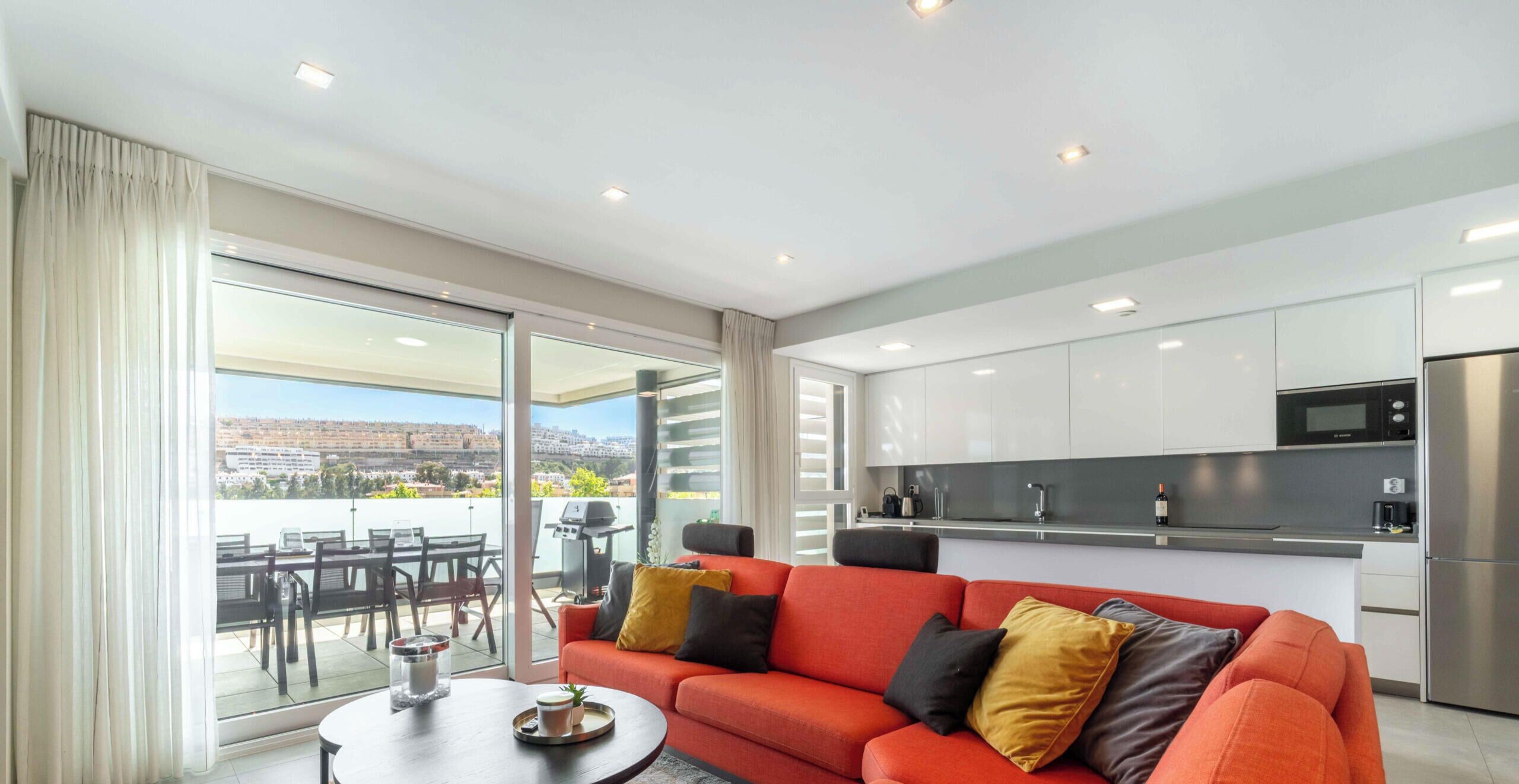 A modern living room with a red sofa, a dining area with a table and chairs, a kitchen with stainless steel appliances, and a sliding glass door leading to a balcony with a view of the city.