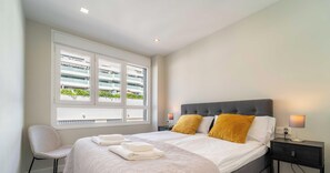 Ein Hotelzimmer mit Bett, zwei Nachttischlampen, einem Sessel, einem Fenster mit Blick auf ein Gebäude und einem Nachttisch.