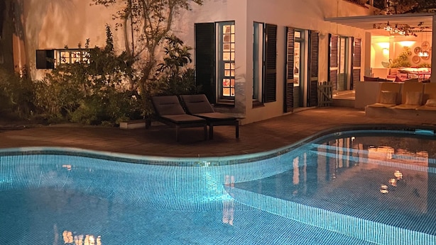 Piscina com borda em azulejo, edifício com fachada branca e persianas verdes, e área de varanda com cadeiras de descanso.