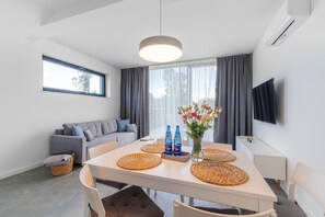 A modern living room with a sofa, a dining table with placemats, a vase with flowers, and a flat-screen TV mounted on the wall.