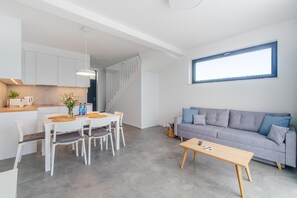 A modern kitchen with white cabinets, a dining area with a table and chairs, a grey sofa, and a large window.