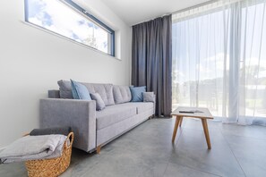 A modern living room with a grey sofa, a small wooden table, a wicker basket, and a large window with curtains. - New holiday homes, Dziwnow (Dziwnów)