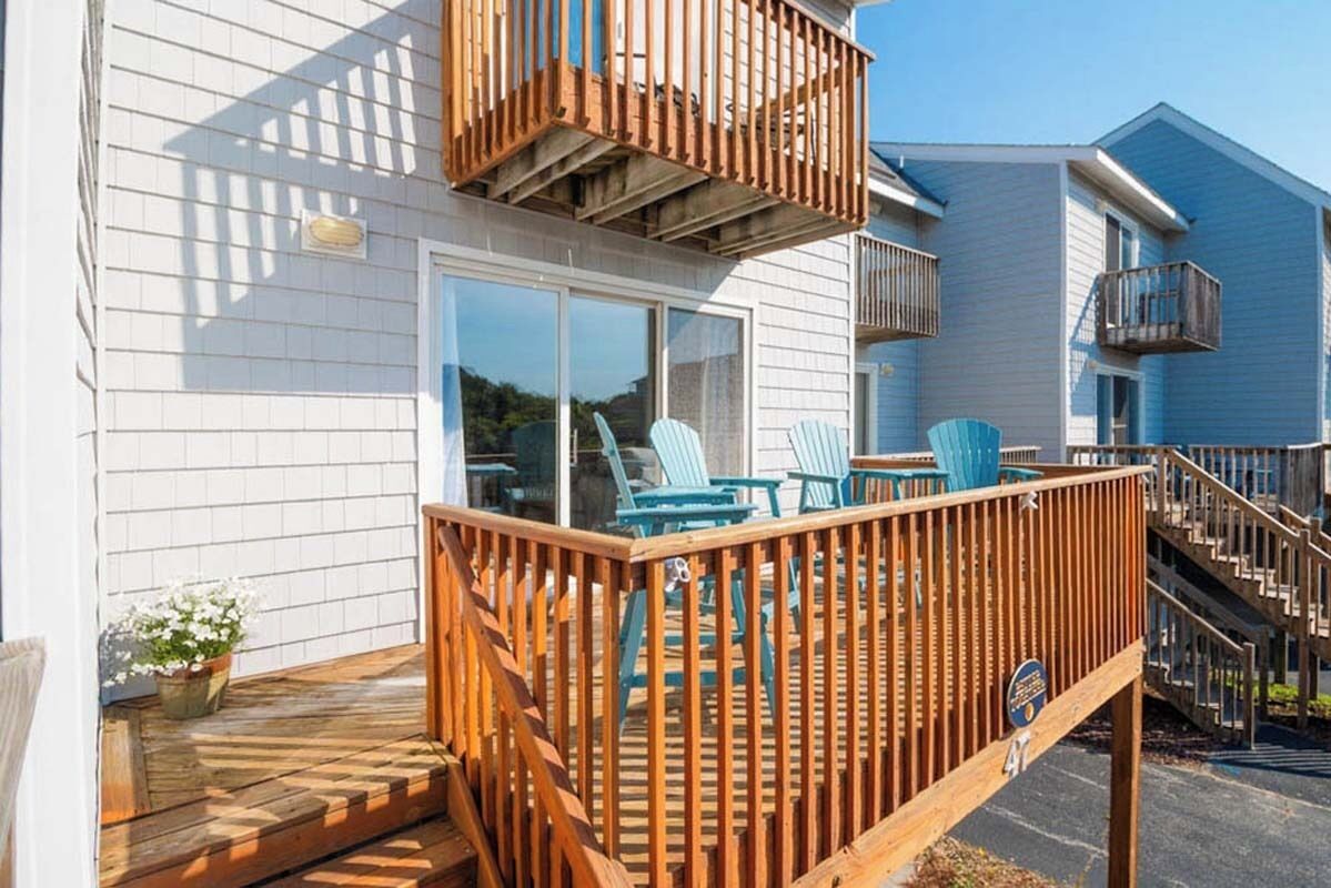 A wooden deck with chairs, a staircase, and a building with white siding and a glass door.
