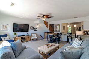 A living room with a TV, sofa, ottoman, and a ceiling fan. - Sun Realty oceanside townhome w/ community pool & beach access (Duck)