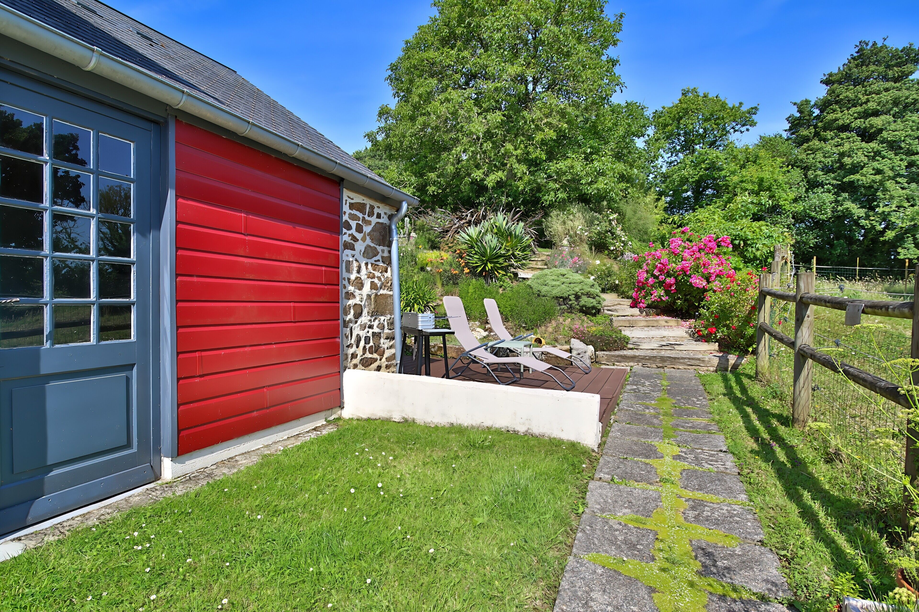 A red building with a stone facade, a stone pathway, and a garden with a wooden fence.