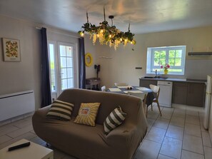 A living room with a sofa, a dining table, a kitchen area, and a ceiling light fixture. - Holiday Home “Le Gîte d’Augustin” with Private Terrace, Garden & Wi-Fi (Sortosville-en-Beaumont)