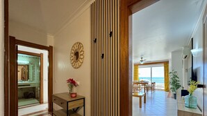 Un couloir mène à une salle à manger offrant une vue sur l’océan. On y trouve un buffet en bois orné d’un vase de fleurs et un miroir accroché au mur.