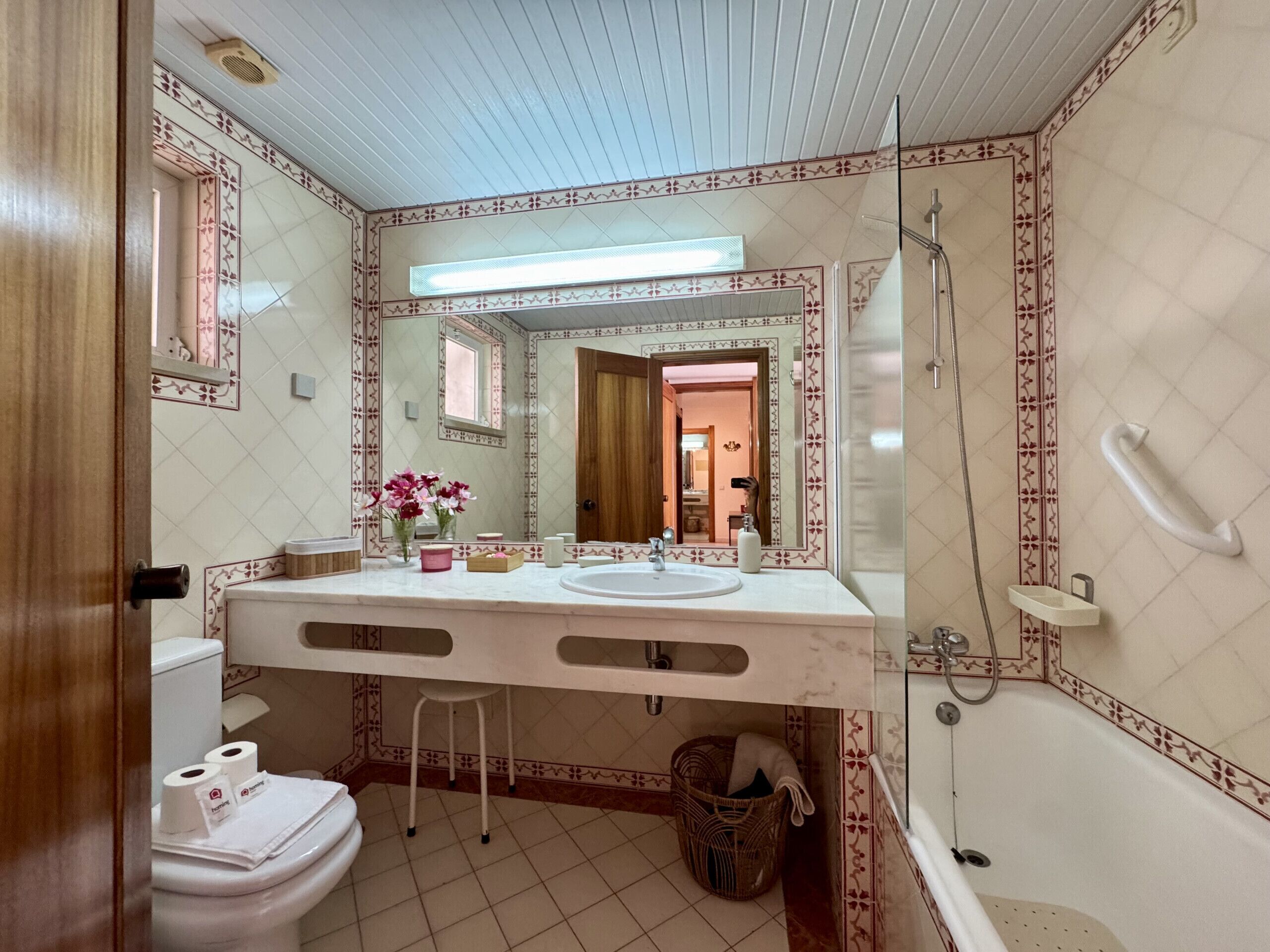 Ein Badezimmer mit Toilette, Waschbecken, Spiegel, Dusche und einem Korbwannenstuhl.