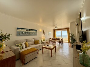 A living room with a sofa, coffee table, and a painting of ducks on water. There's a dining area with a table and chairs, and a view of the sea through a large window. - ALBUFEIRA AMAZING VIEW 2 WITH POOL by HOMING (Albufeira)