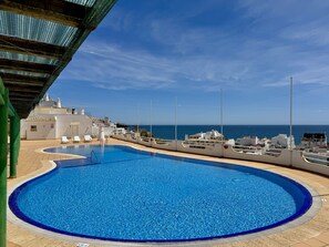 A swimming pool with a clear view of the sea, a covered area with lounge chairs, and a building with a white facade. - ALBUFEIRA AMAZING VIEW 2 WITH POOL by HOMING (Albufeira)