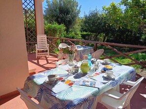 Un'area pranzo all'aperto con un tavolo imbandito per due persone, circondata da sedie, un parapetto e vegetazione sullo sfondo.