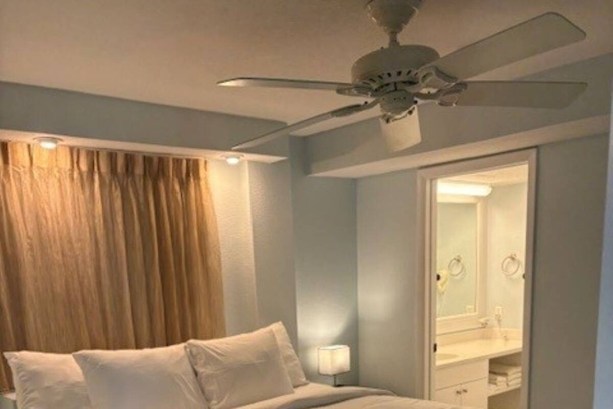 A hotel room with a bed, a ceiling fan, a nightstand, a mirror, and a bathroom visible through an open door.