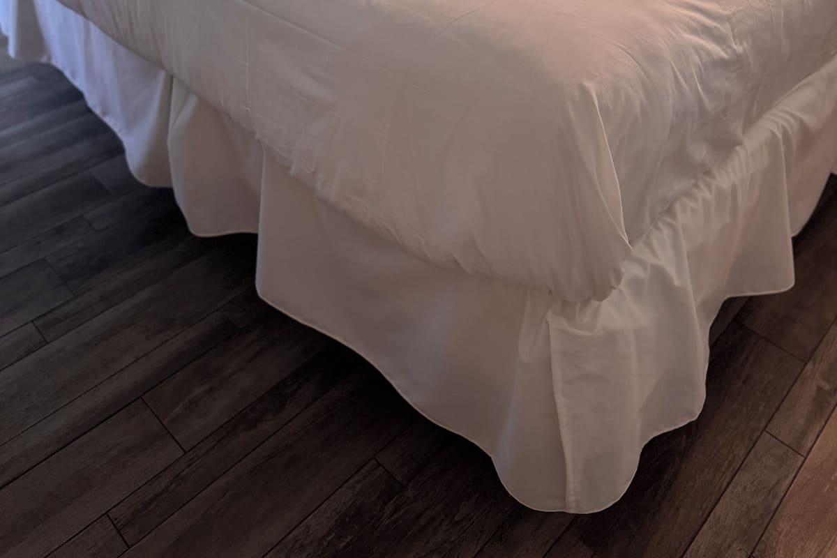 A double bed with white bedding, a small bedside table, a wall-mounted shelf with a decorative item, and a wooden floor.