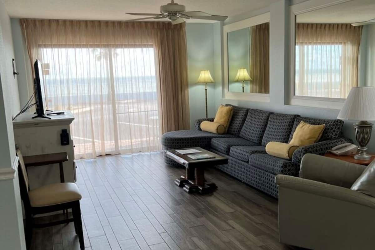 A living room with a sofa, armchair, and a coffee table. There is a ceiling fan, a TV, and a window with curtains.
