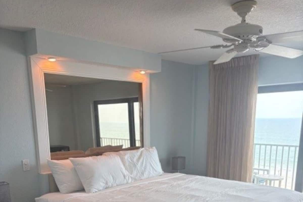 A hotel room with a large bed, a ceiling fan, a nightstand, and a view of the sea.