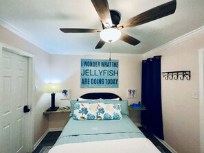 Un dormitorio con ventilador de techo, una cama con almohadas estampadas, un reloj de pared y un cartel con una cita sobre medusas.