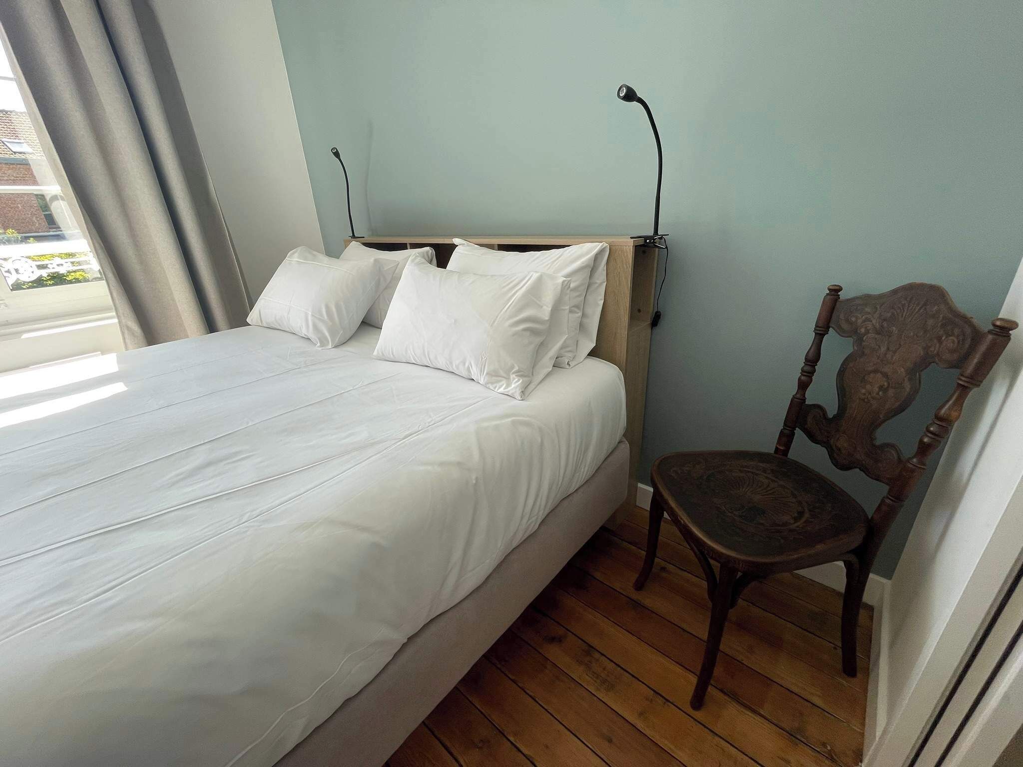 A bed with white linens, a wooden headboard, a chair, and a window with curtains.