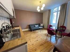 A modern living space with a kitchen area, a sofa, a dining table with chairs, and a large window with curtains. - Queen Bed • Fully Equipped Apartment • Washing Machine • Amiens Centre • 5-min Walk to Station • Parking on Site (Amiens)