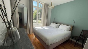 Quarto com cama, um vaso com galhos, janela com cortinas e uma cadeira.