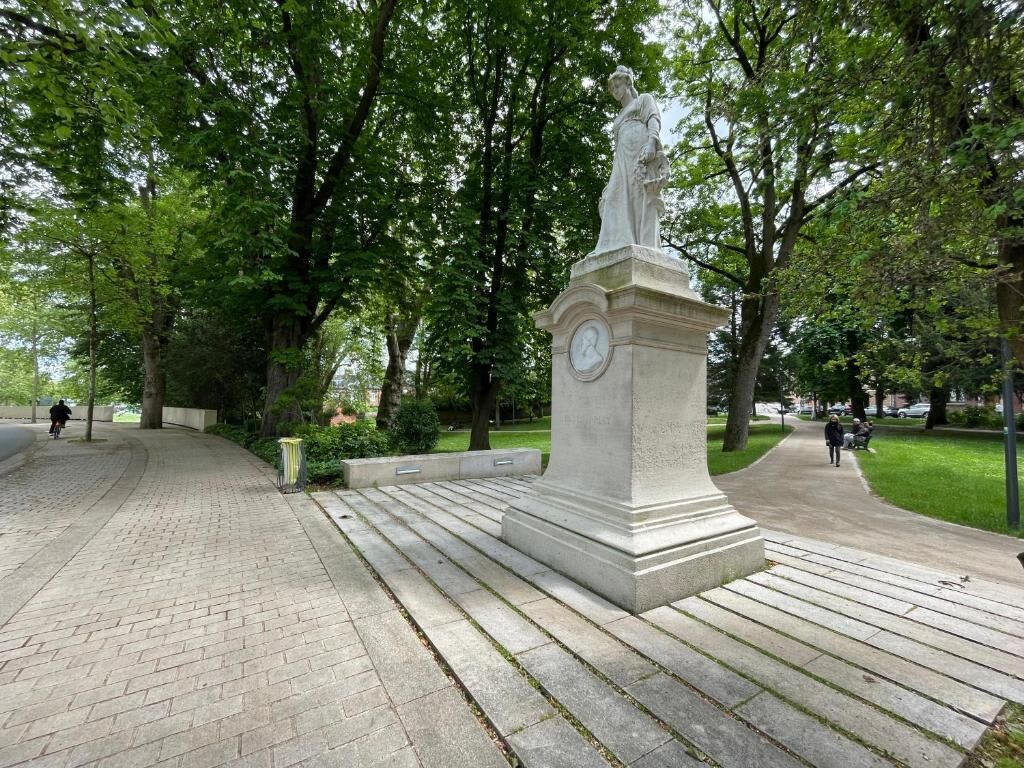 Une statue dans un parc, surmontée d’une horloge, entourée d’arbres et bordée d’une allée pavée.